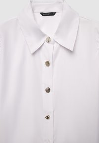 Camicia bianca con colletto a punta, caratterizzata da bottoni argentati texturizzati lungo il davanti e una superficie in tessuto liscia.