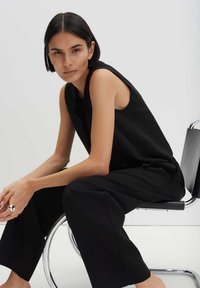 Young person with short dark hair wearing sleeveless black top and pants, sitting sideways on a modern metal-framed chair.