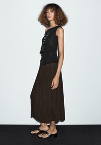 Donna con capelli ricci che indossa un top senza maniche nero, una gonna plissettata marrone, sandali con tacco neri e una lunga collana di perline, in piedi di lato.