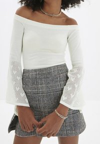 Weißes Off-Shoulder-Oberteil mit ausladenden Netzärmeln, die Herzmuster zeigen, kombiniert mit einem grauen Karorock.