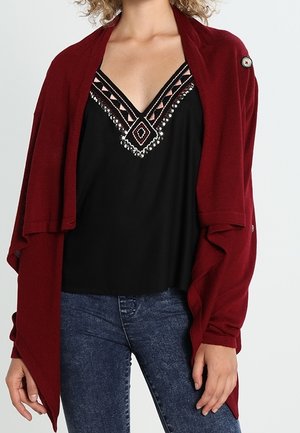 Femme portant un cardigan bordeaux à ouverture frontale sur un haut noir sans manches avec un col géométrique orné de perles et un jean bleu.