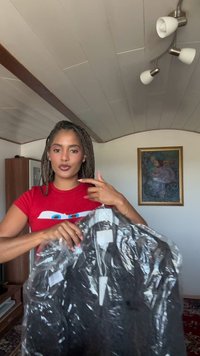 Črna jakna v prozorni torbi, ki prikazuje gladko teksturo in jasno vidne oznake. Model nosi rdečo majico z grafičnim dizajnom.