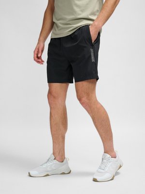 Mand iført sorte Hummel-shorts, beige t-shirt og hvide sneakers står mod en ensartet lys grå baggrund.
