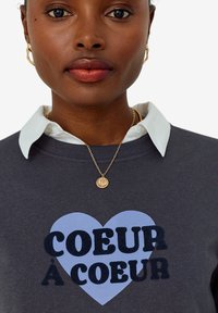 Gray sweatshirt with a blue heart graphic and text "COEUR À COEUR" in black. Features a white collared shirt beneath and a gold pendant necklace.