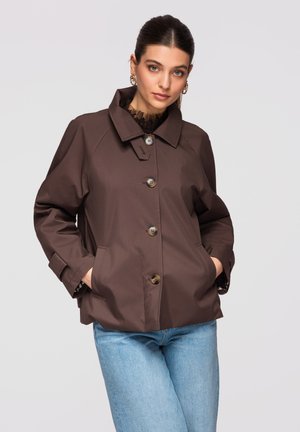 Femme portant une veste boutonnée marron foncé avec col et un jean bleu clair, les mains dans les poches, sur un fond uni gris clair.