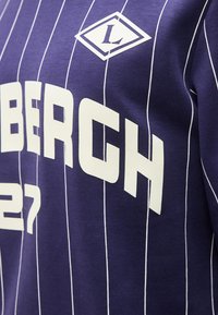 Κοντινή λήψη πορφυρού πουκαμίσου με λευκές κάθετες ρίγες και έντονους λευκούς χαρακτήρες που σχηματίζουν εν μέρει τη λέξη «BERGH» και λογότυπο σε σχήμα ρόμβου με το γράμμα «L».