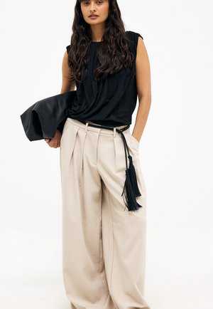 Donna con lunghi capelli scuri che indossa un top nero senza maniche e pantaloni beige larghi con cintura nera con nappine, mentre tiene una giacca nera.