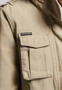 Khakifarvet jakke med en markant brystlomme og en sort label. Fremstillet af robust stof med en glat tekstur og struktureret design.