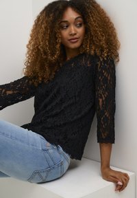 Black lace top with three-quarter sleeves, floral pattern, scalloped edges, paired with light blue jeans. Sitting against a neutral background.