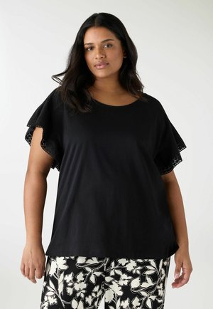 Donna con capelli lunghi e scuri che indossa una maglia nera con maniche bordate di pizzo e pantaloni a fantasia floreale bianco e nera, in piedi davanti a uno sfondo bianco.