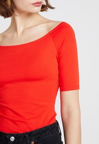 Haut rouge ajusté avec un large décolleté carré et des manches courtes, fabriqué en tissu lisse. Le vêtement est porté avec un pantalon noir taille haute.