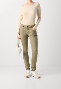 Lichtbeige top met lange mouwen gecombineerd met olijfgroene slim-fit broek. Witte sneakers met lichtgrijze accenten. Houdt een crème kleurige handtas vast.