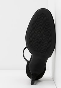 Chaussure à talon haut noire avec semelle texturée et bride à la cheville, inclinée pour montrer la semelle extérieure sur fond blanc à coins.