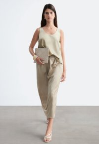 Femme marchant en haut beige sans manches et pantalon ample, tenant une pochette tissée, portant des boucles d'oreilles en or, un bracelet, des bagues et des sandales à lanières.