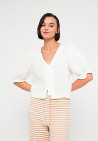 Λευκό πουκάμισο από λινό με κοντά μανίκια, V-λαιμόκοψη, κουμπιά στο μπροστινό μέρος και βολάν στις μανσέτες· φορεμένο με μπεζ και λευκές ριγέ παντελόνες.