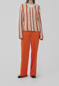 Häkel-Cardigan mit abwechselnd orangefarbenen und cremefarbenen Streifen, ausgestattet mit großen Knöpfen, kombiniert mit hoch geschnittenen orangenen Hosen und schwarzen Schuhen.