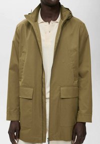Chaqueta impermeable verde oliva con capucha, cierre frontal de cremallera, dos grandes bolsillos y una textura suave, llevada sobre una camisa de color claro.