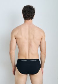 Schwarze Baumwollslips mit einer glatten Textur, ausgestattet mit einem gebrandeten Bund und einer engen Passform, von hinten betrachtet.
