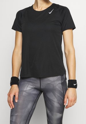 Person trägt ein schwarzes Nike-Kurzarmshirt, graue gemusterte Leggings und schwarze Nike-Handgelenkbänder und steht vor einem schlichten Hintergrund.