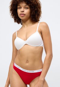 Λευκό σουτιέν με επένδυση και ρυθμιζόμενες τιράντες, συνδυασμένο με κόκκινο thong που φέρει λευκή ζώνη με λογότυπο TOMMY HILFIGER. Απαλή βαμβακερή υφή.