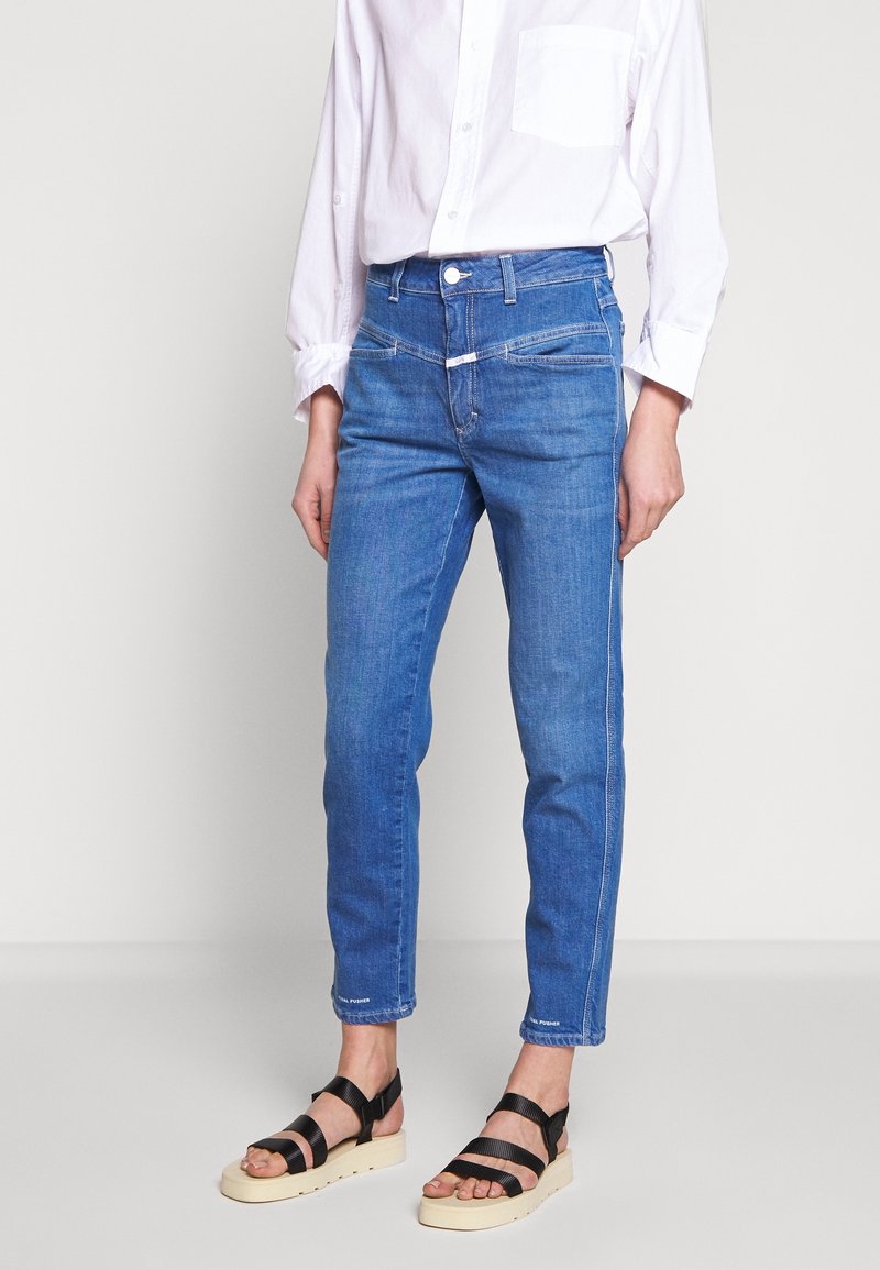 Personne portant un jean bleu taille moyenne, une chemise blanche à manches longues et des sandales à plateforme noires, debout devant un fond clair uni.