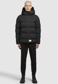 Fekete puffernadrág kapucnival, elöl cipzárral és enyhén fényes textúrával. Fekete nadrággal és csizmával párosítva.