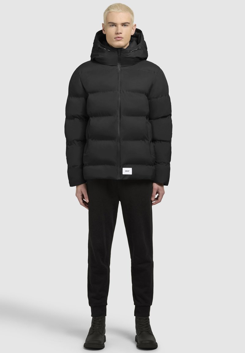 Chaqueta puffer negra con capucha, con cierre de cremallera en el frente y una textura ligeramente brillante. Combinada con pantalones negros y botas.