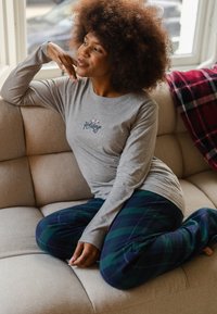 Grijze langemouwshirts met de tekst "Fijne Feestdagen". Gecombineerd met donkere geruite broeken met groene, blauwe en rode strepen. Zittend op een beige bank.