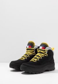 Černé turistické boty se žlutými tkaničkami, svršek z nubuku a síťoviny, polstrovaný límec, gumová podrážka a detail loga na jazyku. Odolný design pro venkovní použití.