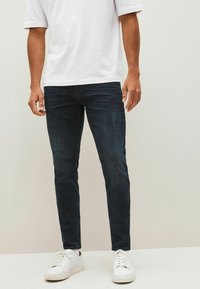 Temno modri jeans iz denima s slim fit krojem, z diskretnim zbledelimi detajli, v kombinaciji z enostavno belo majico in belimi supergami.