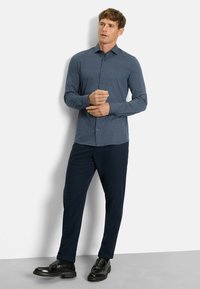 Langarmhemd in Marineblau mit geometrischem Muster, kombiniert mit marineblauen Hosen und schwarzen Lederschuhen. Das Hemd hat einen kragenfreien Hals und Knöpfe.