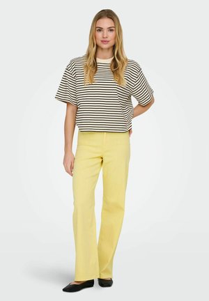 Jeune femme debout, main sur la hanche, portant une chemise ample à rayures noires et blanches, un pantalon jaune et des ballerines noires sur fond blanc.