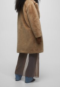 Abrigo de piel sintética en beige, con un corte holgado y mangas largas. Lleva puesto sobre un vestido fluido y transparente con rayas marrones y beige y unos pantalones vaqueros de pierna ancha.