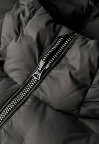 Tejido de chaqueta acolchada negra con textura quilted. Presenta un cierre metálico con una etiqueta "DRY", acentuado por costuras en contraste.