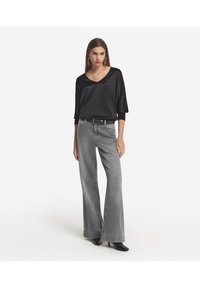 Schwarze Satinbluse mit V-Ausschnitt und drei Viertelärmeln, kombiniert mit grauen weit geschnittenen Jeans und schwarzen Absatzschuhen. Schlichtes Design.