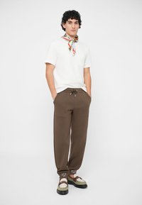 Camicia bianca a maniche corte con tasca, pantaloni marroni con cordino, e sciarpa a fantasia multicolore. Le scarpe combinano dettagli marroni e bianchi.