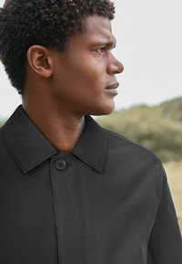 Manteau noir avec un col, fermeture à un seul bouton et texture lisse. Profil du mannequin montrant des cheveux courts et bouclés naturels sur un fond neutre.