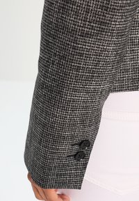 Tissu à carreaux noir et blanc texturé d'une manche de veste sur mesure, avec deux boutons noirs au poignet et une chemise blanche sans couture en dessous.