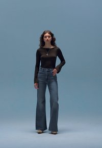 Haut noir à manches longues associé à un jean flare taille haute bleu clair et des talons beiges. Un collier ajoute une touche subtile. Fond bleu clair.