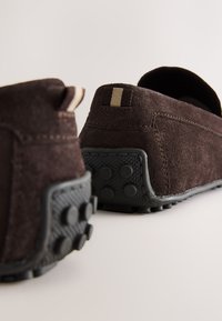 Chaussures en daim marron avec une semelle ruban noir texturée, présentant une étiquette rayée subtile à l’arrière et une forme arrondie pour le confort.