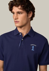 Navy modré polo triko s jemnou texturou, se vyšitým nápisem "HACKETT" a číslem "1" v kontrastní modré barvě, s knoflíkovým límcem a krátkými rukávy.