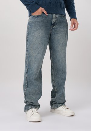 Uomo che indossa jeans blu sbiaditi a vestibilità ampia, scarpe da ginnastica bianche e un maglione blu scuro con mano in tasca, in piedi su sfondo grigio chiaro.