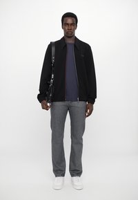 Veste noire zippée avec des accents rouges, chemise bleu marine en dessous, jeans gris et baskets blanches. Modèle tenant un sac à bandoulière noir.