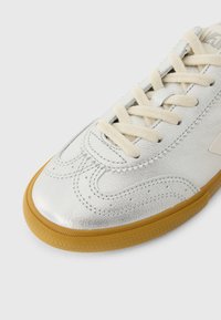 Ασημένιο δερμάτινο αθλητικό παπούτσι με κρέμ κορδόνια, που διαθέτει στρογγυλεμένη μύτη, υφή στην επιφάνεια και σόλα από καουτσούκ γκαμ. Υπάρχει περίτεχνη λεπτομέρεια ραφής.