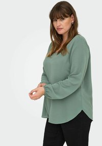Minta zaļa garām piedurknēm blūze ar brīvu piegriezumu, aprīkota ar noapaļotu apakšmalu, elastīgām piedurknēm un teksturētu audumu. Saskaņojot ar melniem biksēm.