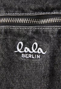 Material de denim preto com um zipper prateado e o logótipo bordado em branco "lala BERLIN" na superfície do tecido, exibindo uma textura suave.