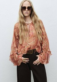 Flowy, gestreepte blouse in warme tinten met uitgerolde mouwen en een striknek, gecombineerd met hooggetailleerde donkere broeken en gouden sieraden.
