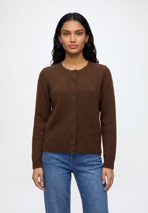 Femme aux longs cheveux foncés, portant un cardigan marron boutonné et un jean bleu, debout devant un fond blanc uni.
