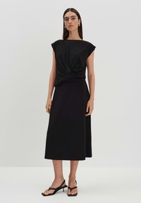 Abito nero con un top corto drappeggiato e dettaglio a torsione, gonna fluente e cinturini eleganti su sandali con tacco nero.