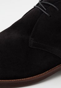 Scarpa in suede nero con lacci, dalla texture liscia, punta rotonda e suola marrone a contrasto con un accento cucito.